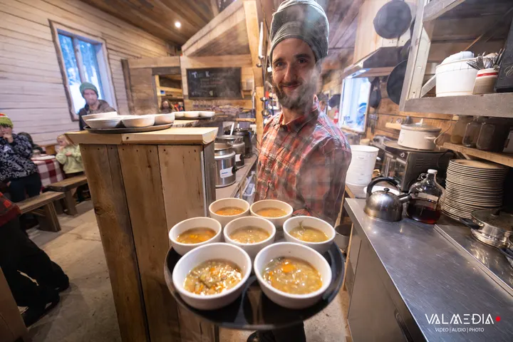 Un serveur souriant porte un plateau de six bols de soupe fumante dans la salle à manger de la cabane
